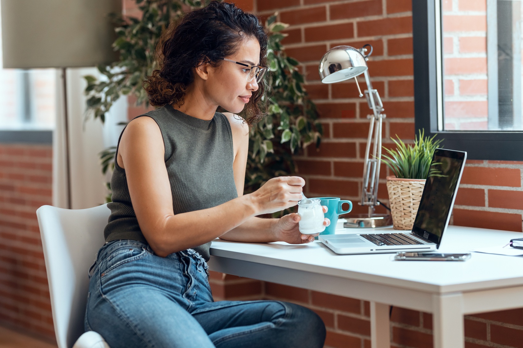 Beautiful young business woman working with laptop while eating yogurt in living room at home.