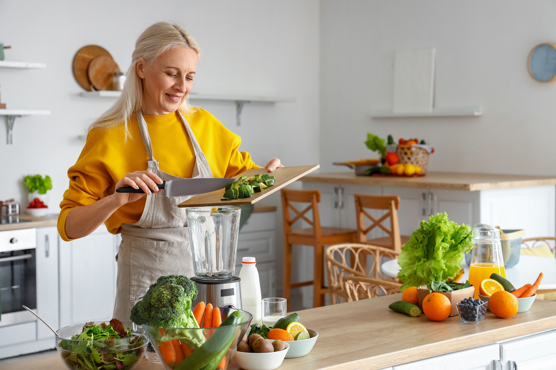 Mature woman with cut avocado making healthy smoothie in kitchen